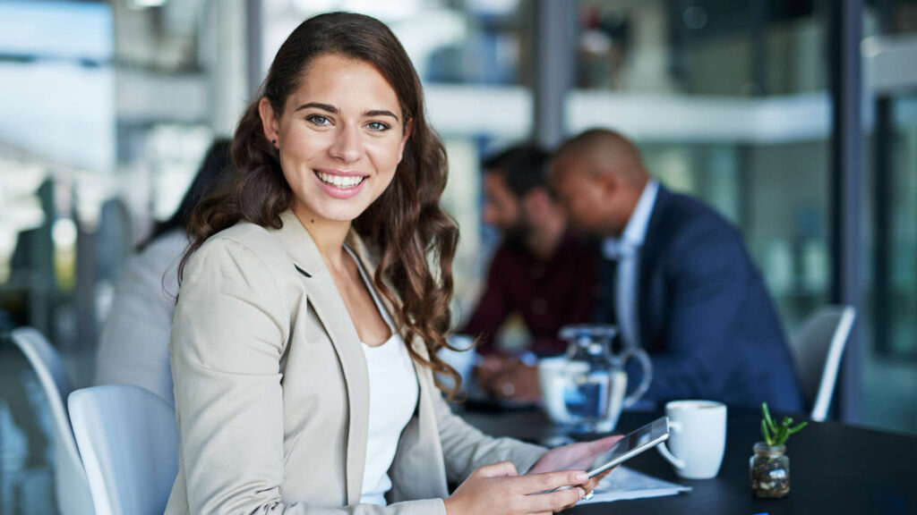 Uśmiechnięta kobieta w elegancko wyposażonym biurze trzyma tablet, gdzie zarówno sprzęt, jak i meble stanowią rzeczowe aktywa trwałe w Holandii – materiële vaste activa.